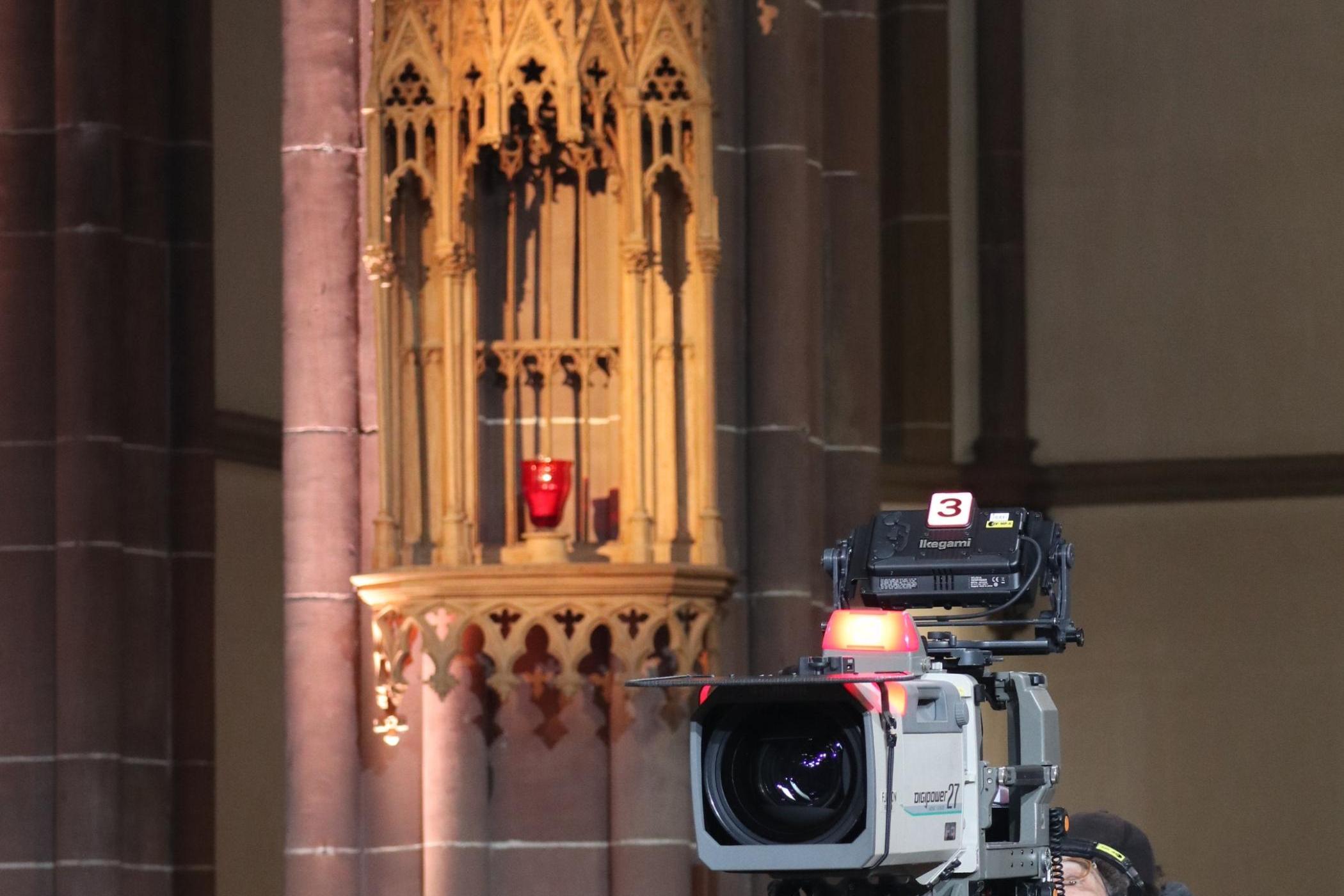In einer Kirche steht eine große Fernsehkamera. Das Aufnahmelicht leuchtet.